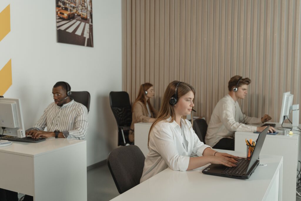 Customer support team at their desks in headsets, an agent typing on a laptop mid-call while colleagues handle their own calls behind her.