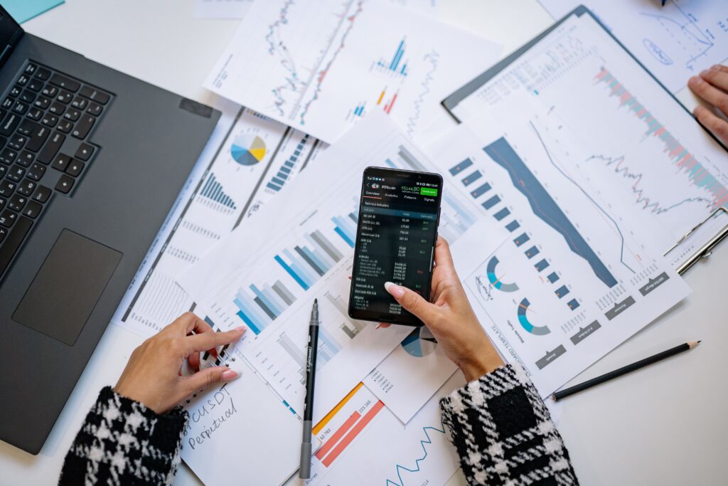 Overhead view of a finance review, hands working through printed charts and reports beside a laptop while checking figures on a phone.