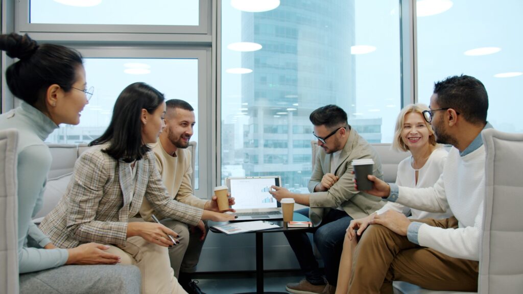 Six colleagues around a laptop in an office lounge, coffees in hand, talking through a chart on the screen together.