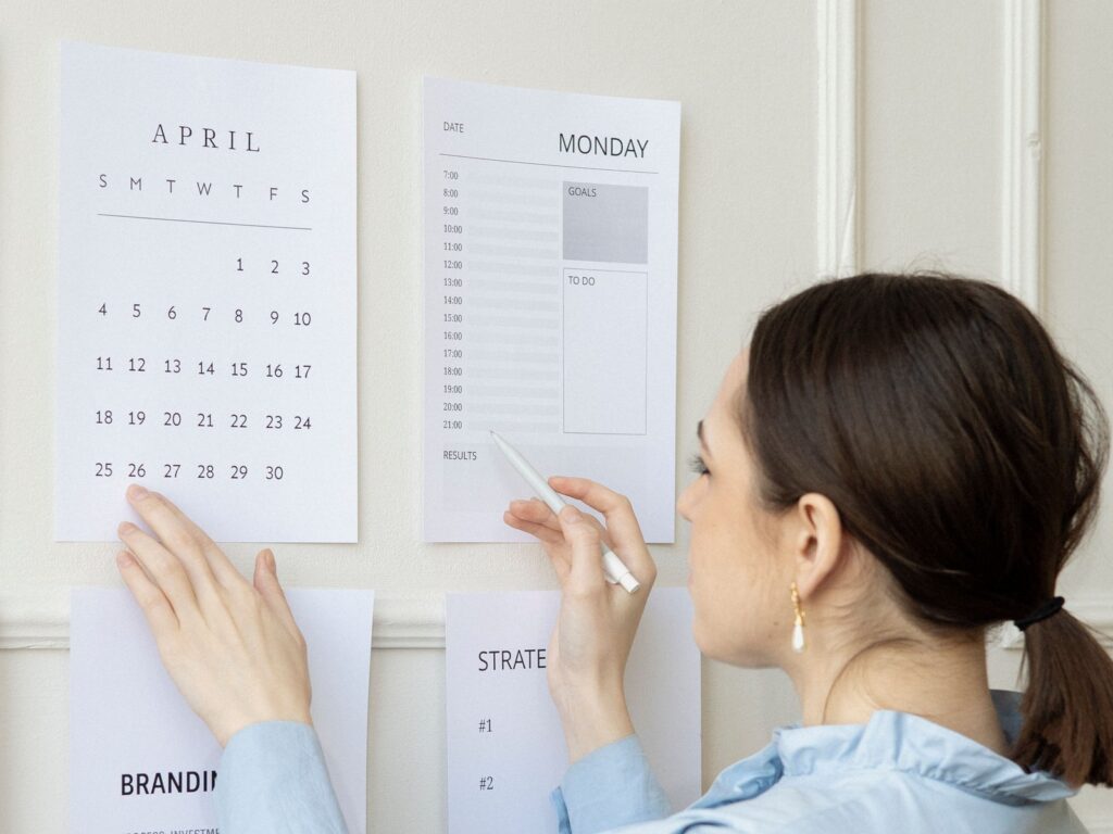 Woman pinning a monthly calendar to the wall with one hand while she marks the day's schedule with a pen in the other.