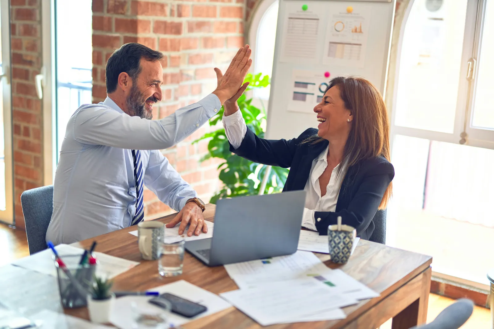 Two colleagues at a meeting table high-five over a laptop and printed charts, both laughing.